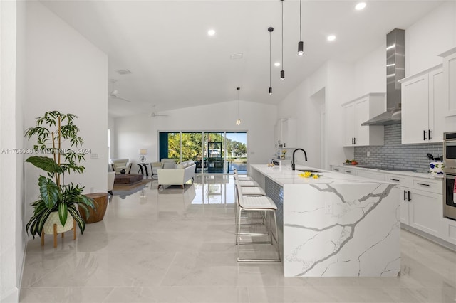 kitchen with wall chimney range hood, light stone counters, a large island with sink, pendant lighting, and lofted ceiling