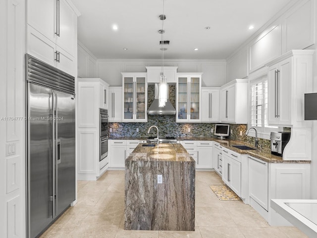 kitchen featuring appliances with stainless steel finishes, dark stone counters, wall chimney exhaust hood, sink, and a center island with sink