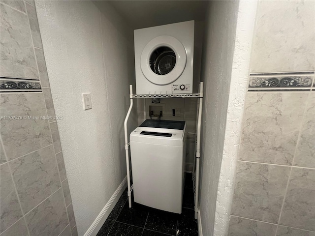 washroom featuring stacked washing maching and dryer, a textured wall, dark tile patterned flooring, and laundry area