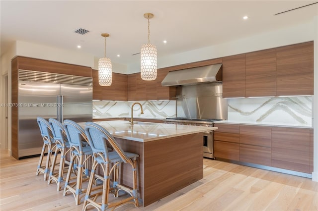kitchen featuring visible vents, a sink, wall chimney range hood, high quality appliances, and brown cabinetry