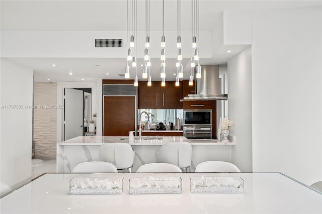 kitchen featuring wall chimney range hood, built in appliances, hanging light fixtures, and sink