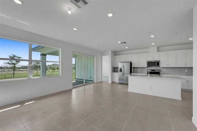 kitchen with light tile patterned floors, decorative backsplash, a center island with sink, white cabinets, and appliances with stainless steel finishes