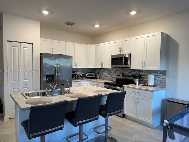 kitchen featuring white cabinets, a center island with sink, decorative backsplash, light tile patterned floors, and stainless steel appliances