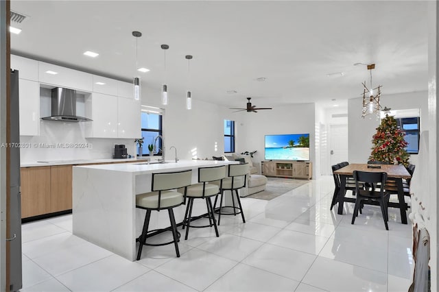 kitchen featuring white cabinets, an island with sink, hanging light fixtures, and wall chimney range hood