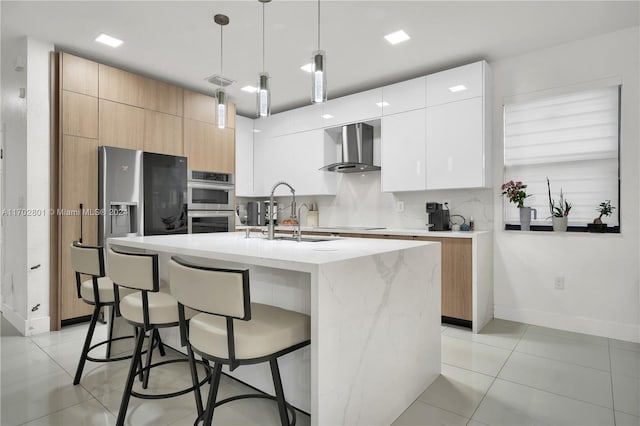 kitchen featuring a kitchen island with sink, white cabinets, wall chimney range hood, sink, and decorative light fixtures