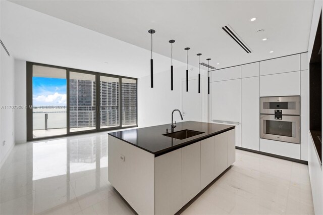 kitchen with floor to ceiling windows, a kitchen island with sink, sink, pendant lighting, and white cabinetry