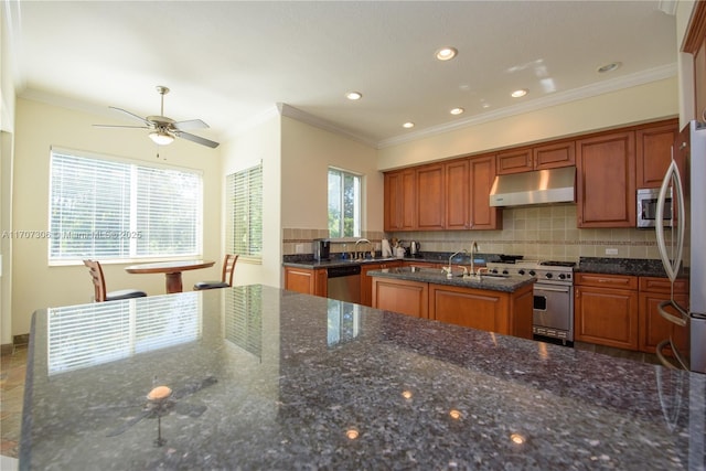 kitchen featuring appliances with stainless steel finishes, an island with sink, decorative backsplash, dark stone counters, and crown molding