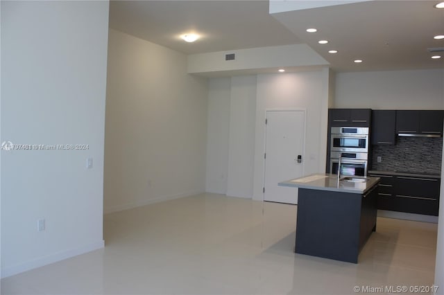 kitchen with double oven, tasteful backsplash, a center island with sink, and light tile patterned floors