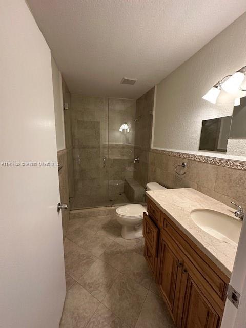 bathroom featuring a textured ceiling, toilet, vanity, a shower with shower door, and tile walls