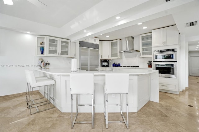 kitchen featuring white cabinets, wall chimney range hood, kitchen peninsula, a breakfast bar area, and stainless steel appliances