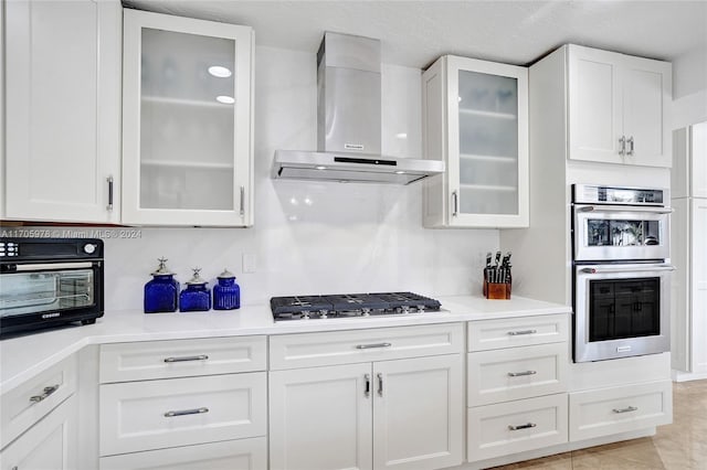 kitchen featuring decorative backsplash, appliances with stainless steel finishes, wall chimney exhaust hood, light tile patterned floors, and white cabinetry