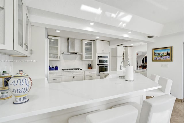 kitchen with white cabinetry, sink, wall chimney range hood, kitchen peninsula, and a kitchen bar