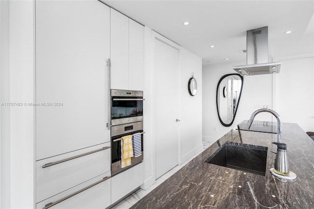 kitchen with white cabinets, double oven, island exhaust hood, and dark stone countertops