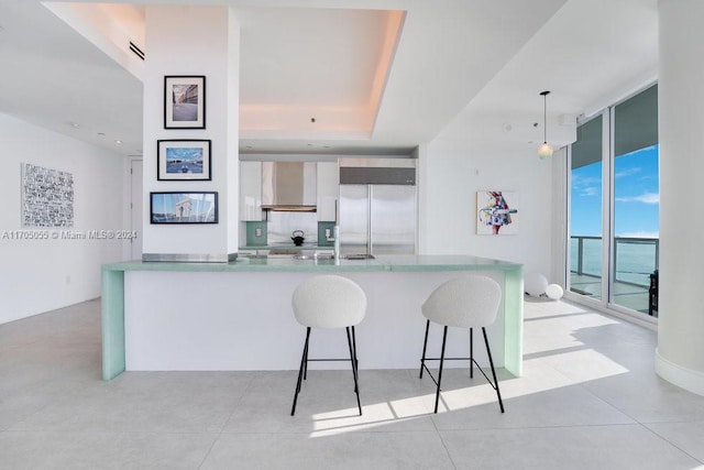 kitchen with wall chimney exhaust hood, a wall of windows, built in fridge, pendant lighting, and a breakfast bar