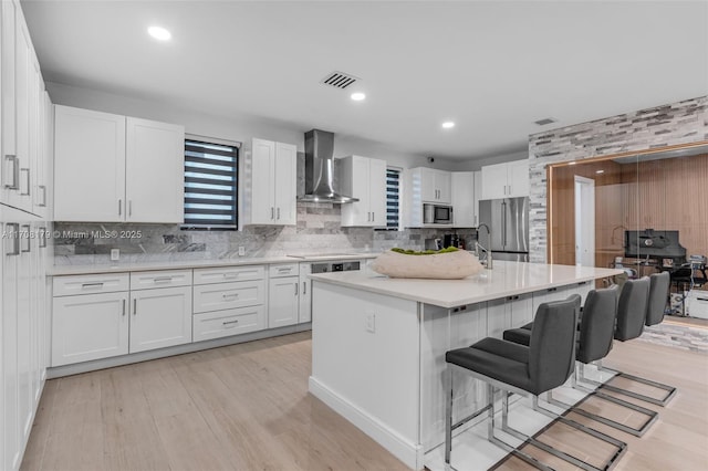 kitchen featuring wall chimney range hood, a breakfast bar, stainless steel appliances, white cabinets, and a center island with sink