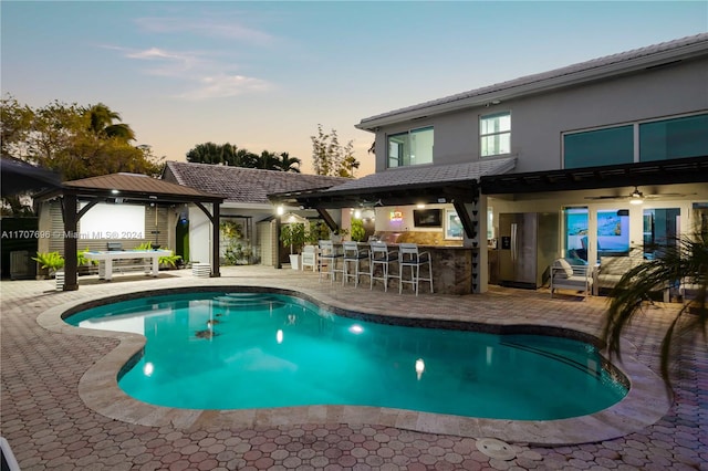 pool at dusk with a gazebo, a bar, and a patio