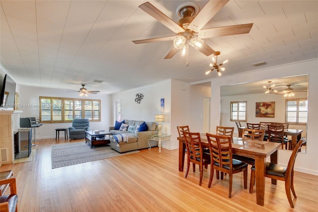 dining space featuring a tiled fireplace, crown molding, light hardwood / wood-style floors, and ceiling fan with notable chandelier