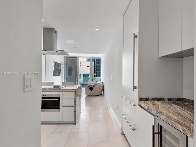 kitchen with black electric stovetop, wall chimney exhaust hood, oven, white cabinets, and wine cooler