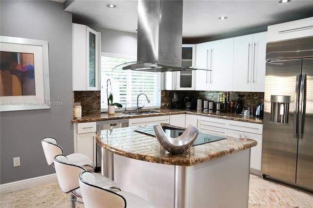 kitchen with sink, stainless steel appliances, dark stone countertops, island range hood, and white cabinets