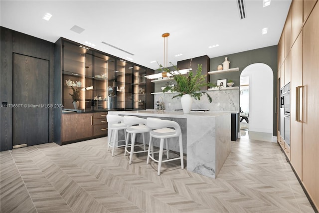 kitchen with light stone countertops, light parquet floors, an island with sink, decorative light fixtures, and a breakfast bar