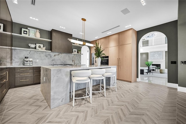 kitchen with backsplash, a kitchen breakfast bar, hanging light fixtures, an island with sink, and light parquet flooring