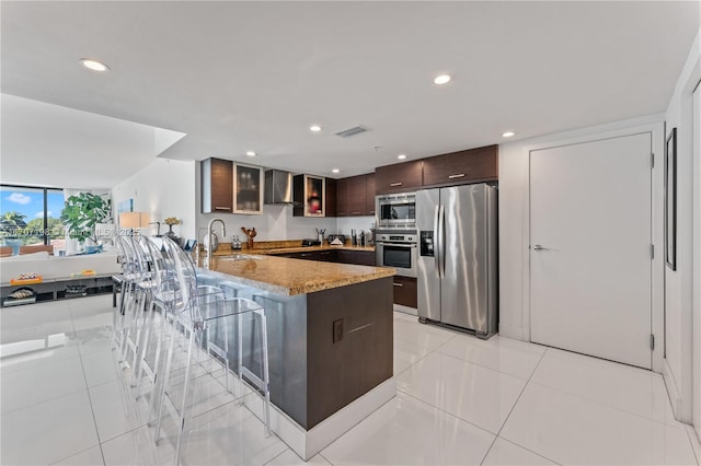 kitchen with a peninsula, dark brown cabinetry, wall chimney range hood, a sink, and stainless steel appliances
