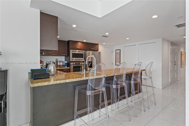 kitchen with light tile patterned floors, light stone counters, appliances with stainless steel finishes, a kitchen bar, and kitchen peninsula