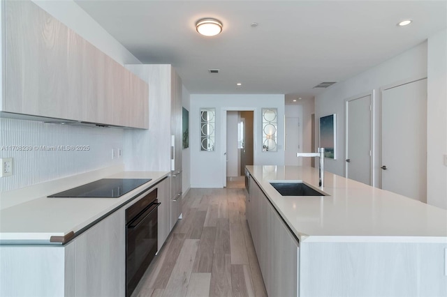 kitchen featuring black appliances, a center island with sink, sink, light hardwood / wood-style flooring, and light brown cabinetry