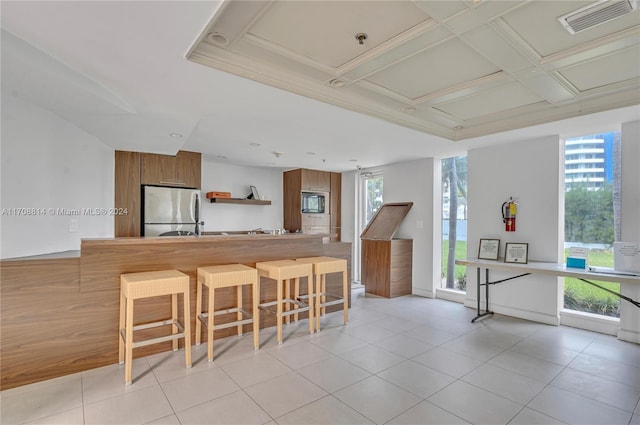 kitchen featuring coffered ceiling, stainless steel fridge, light tile patterned floors, built in microwave, and a kitchen bar