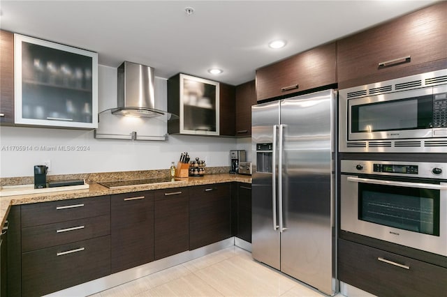 kitchen with light stone counters, dark brown cabinetry, stainless steel appliances, wall chimney range hood, and light tile patterned floors