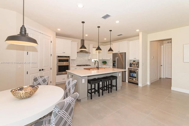 kitchen with white cabinetry, stainless steel appliances, hanging light fixtures, wall chimney range hood, and an island with sink