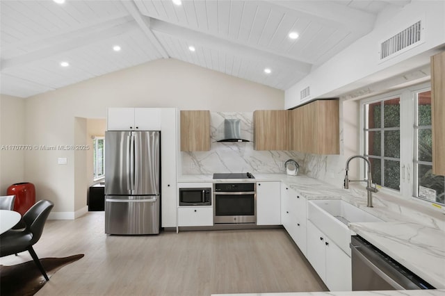 kitchen with appliances with stainless steel finishes, lofted ceiling with beams, sink, white cabinets, and wall chimney range hood