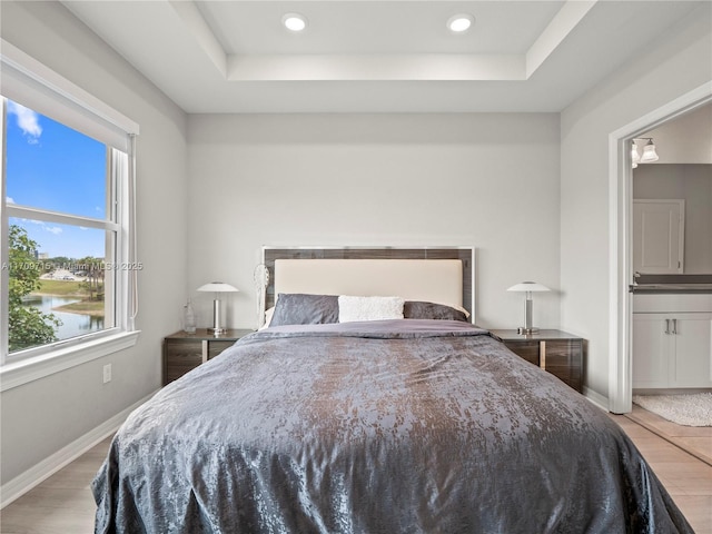 bedroom featuring hardwood / wood-style flooring, a water view, connected bathroom, and a raised ceiling