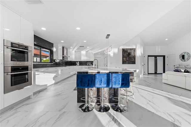 kitchen with a kitchen breakfast bar, stainless steel double oven, pendant lighting, a center island with sink, and white cabinets