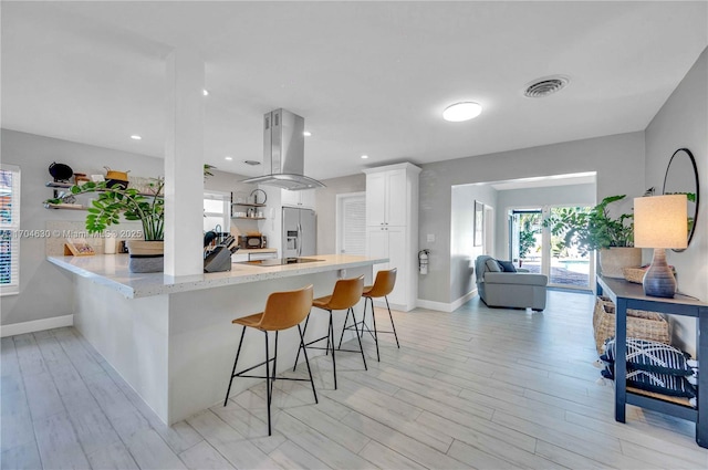 kitchen with white cabinetry, stainless steel refrigerator with ice dispenser, kitchen peninsula, island exhaust hood, and a breakfast bar area