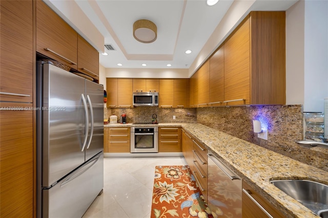 kitchen featuring sink, light stone counters, appliances with stainless steel finishes, a raised ceiling, and decorative backsplash