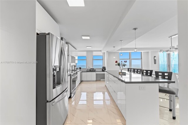 kitchen featuring wall chimney exhaust hood, stainless steel appliances, white cabinets, decorative light fixtures, and a water view