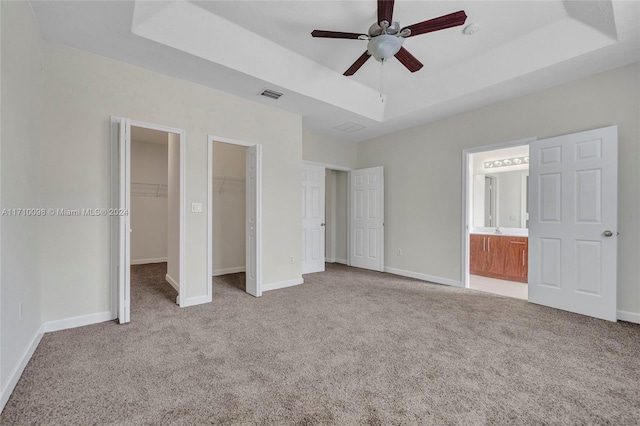 unfurnished bedroom with light carpet, ensuite bath, a tray ceiling, ceiling fan, and a spacious closet