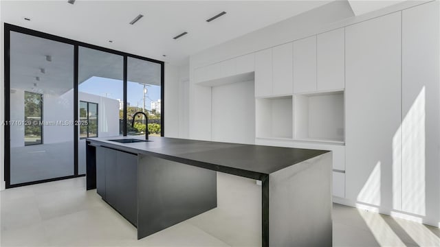 kitchen with a wall of windows, white cabinetry, a kitchen island with sink, and sink