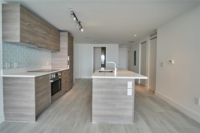 kitchen with tasteful backsplash, sink, rail lighting, a kitchen island with sink, and oven