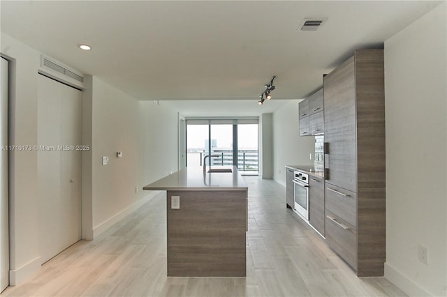 kitchen with stainless steel oven, a kitchen island with sink, light wood-type flooring, track lighting, and sink