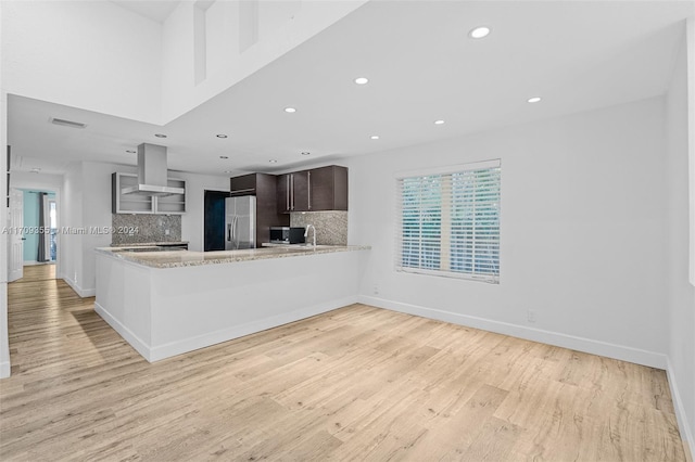 kitchen with dark brown cabinetry, wall chimney range hood, tasteful backsplash, kitchen peninsula, and appliances with stainless steel finishes