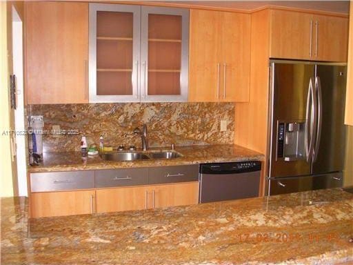 kitchen with stainless steel appliances, a sink, decorative backsplash, glass insert cabinets, and light stone countertops