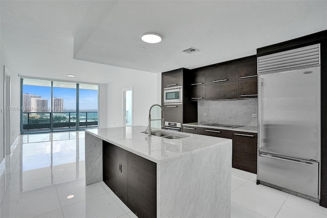 kitchen with sink, built in appliances, light tile patterned flooring, an island with sink, and a wall of windows