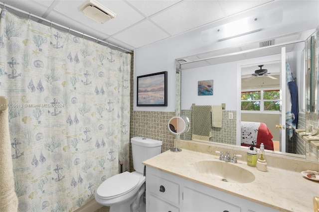 bathroom with a drop ceiling, vanity, ceiling fan, tile walls, and toilet