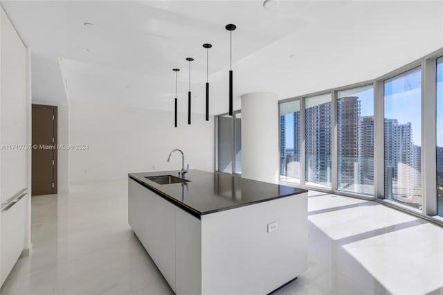 kitchen with sink, hanging light fixtures, a wall of windows, a center island with sink, and white cabinets