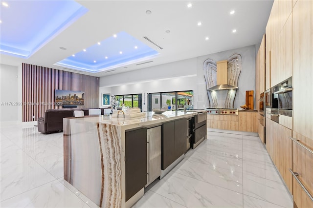 kitchen featuring light stone counters, stainless steel oven, a tray ceiling, a large island with sink, and light brown cabinets