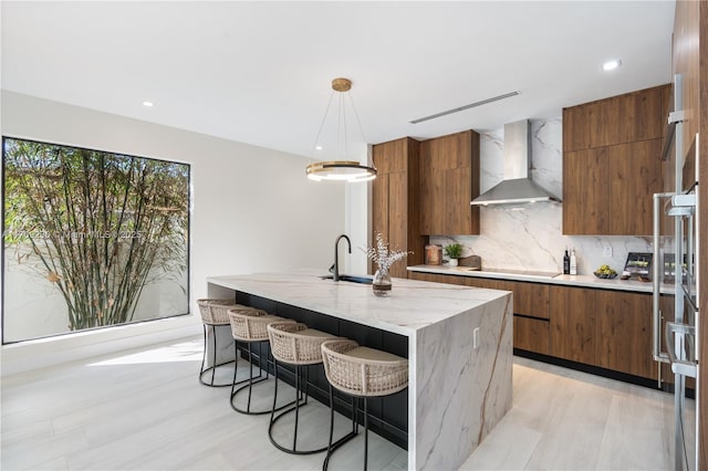 kitchen featuring a breakfast bar, a center island with sink, wall chimney range hood, tasteful backsplash, and decorative light fixtures