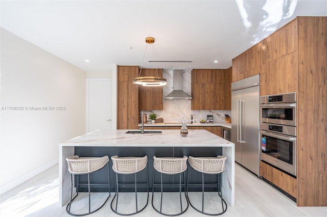 kitchen with stainless steel appliances, wall chimney range hood, a large island with sink, hanging light fixtures, and a breakfast bar area