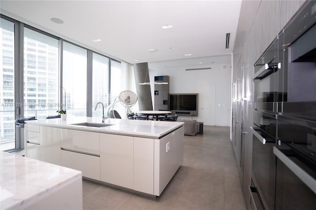 kitchen featuring light stone counters, floor to ceiling windows, sink, white cabinetry, and an island with sink